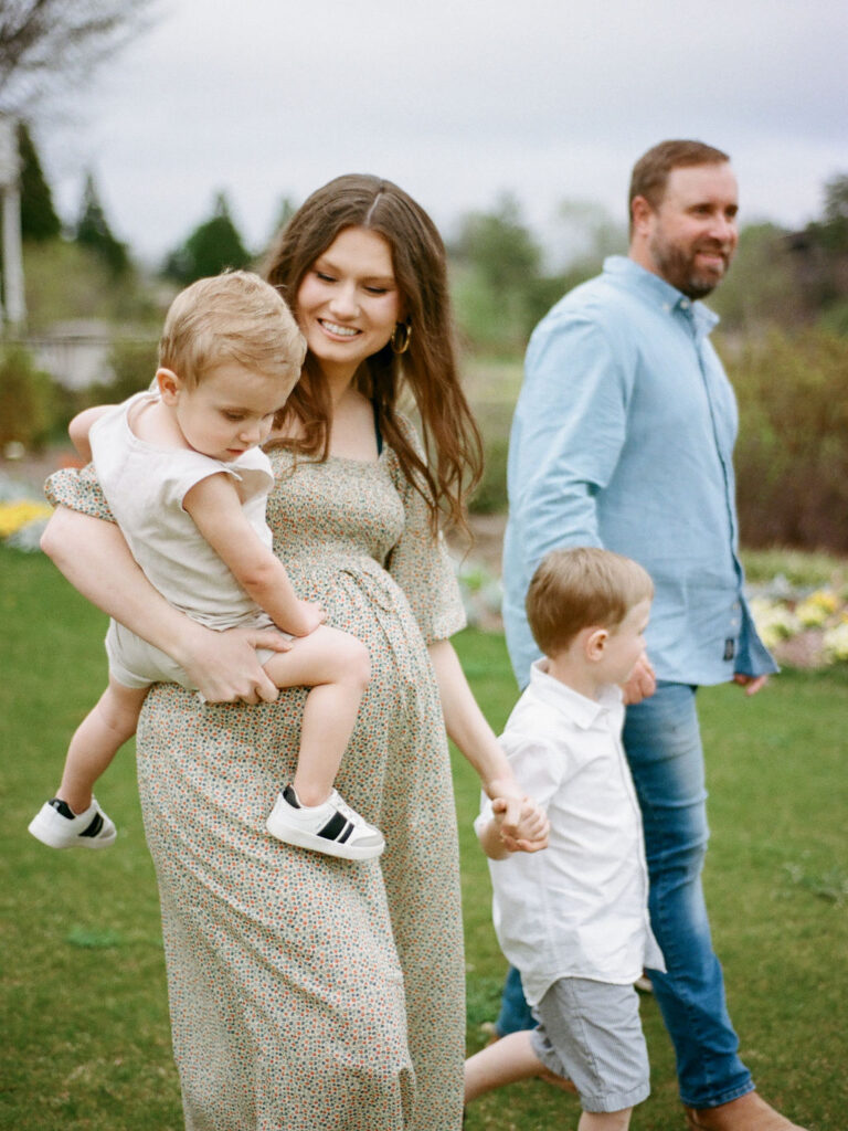 A candid moment of a pregnant mother taking maternity photos with her kids and husband, holding one child on her hip and holding hands with another child.