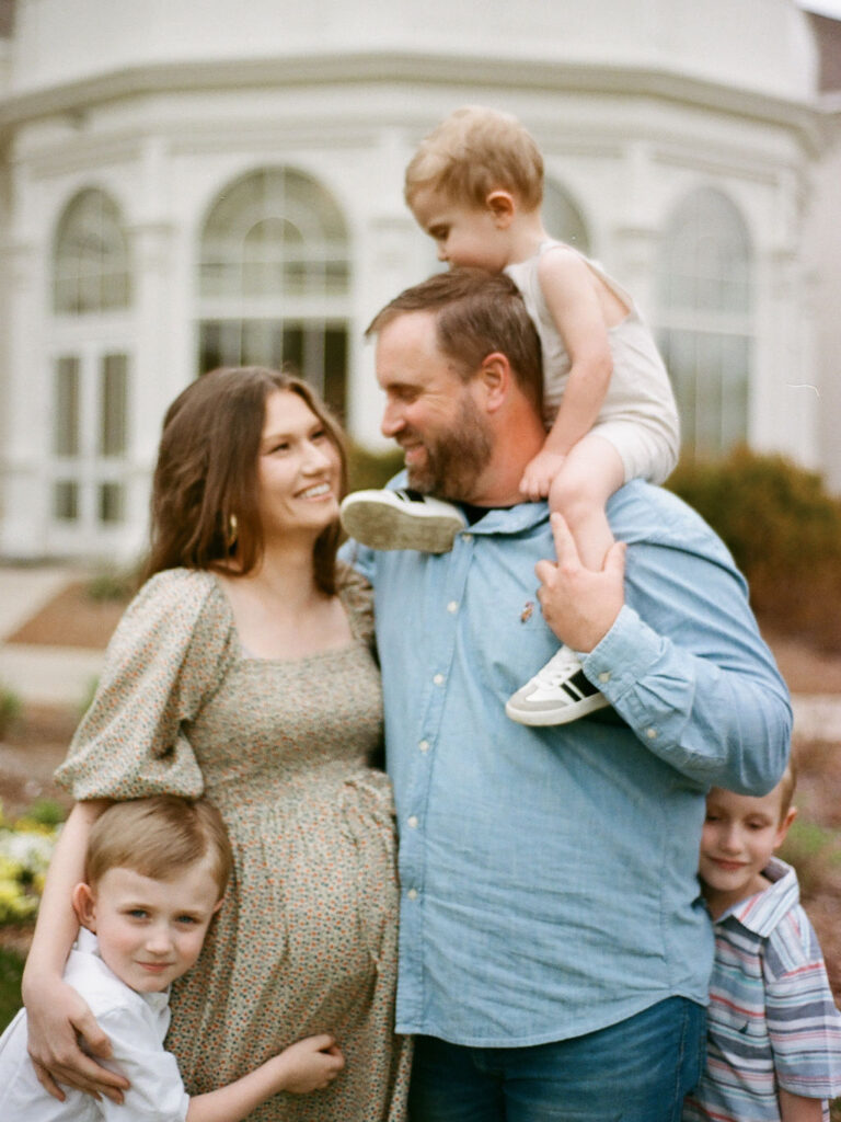 Parents looking at each other with their toddler on dad's shoulders while their two older children hug their legs. Taken on film at the Huntsville Botanical Garden.