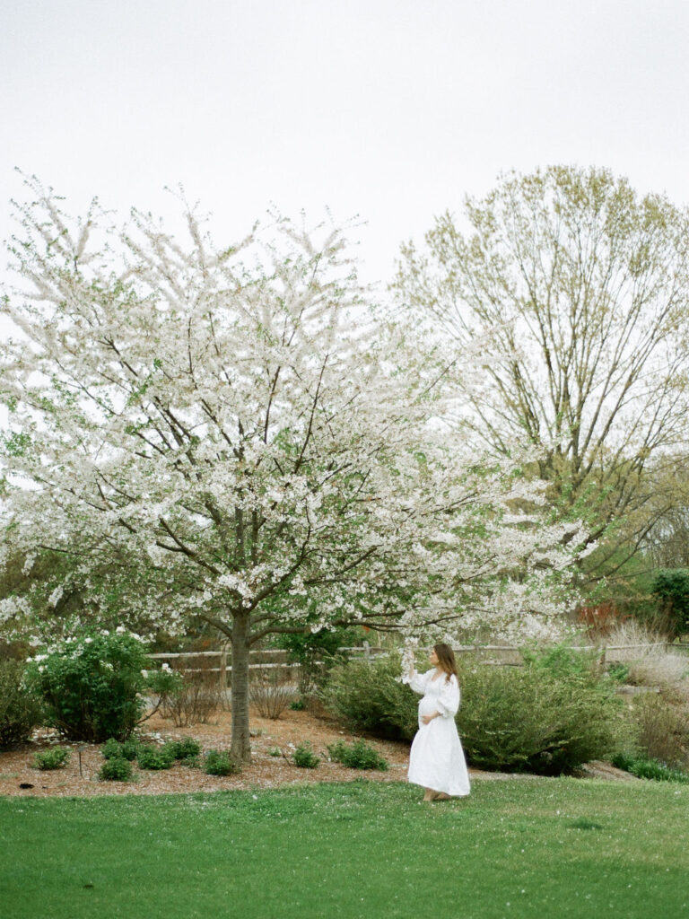 A pregnant woman in a white dress walks through the Huntsville Botanical Garden at her North Alabama maternity photo session.