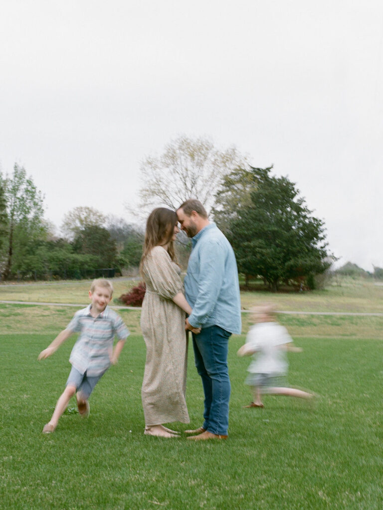 A couple hold hands, facing each other with their foreheads together, while their blurry kids run around them at the Huntsville Botanical Garden.