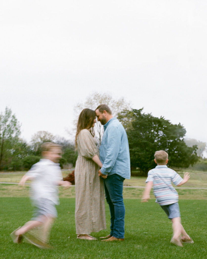 A couple hold hands, facing each other with their foreheads together, while their blurry kids run around them at the Huntsville Botanical Garden.