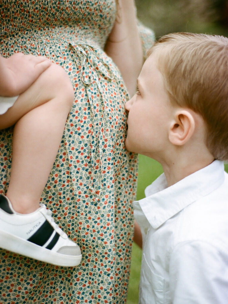 A young child kisses his mother's pregnant belly at her Huntsville Botanical Garden maternity photo session.