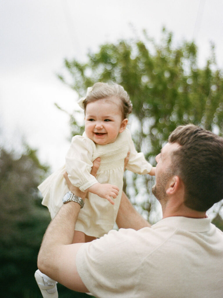 A dad gently boosts his toddler daughter above his head while she laughs at a Huntsville Botanical Garden photography session