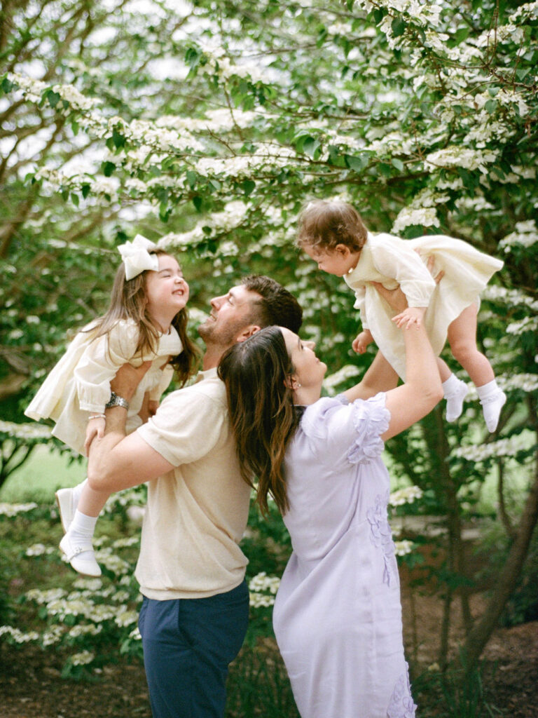 Two parents stand back to back while they each boost one of their daughters above their heads at their Huntsville Botanical Garden family session