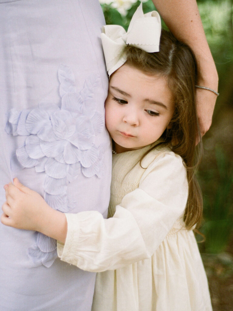 A small child in a yellow dress hugs her mother's leg during their Huntsville Botanical Garden family photoshoot
