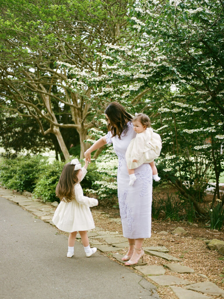 A mother holds her toddler on one hip while her other daughter does a twirl at their Huntsville Botanical Garden family photoshoot