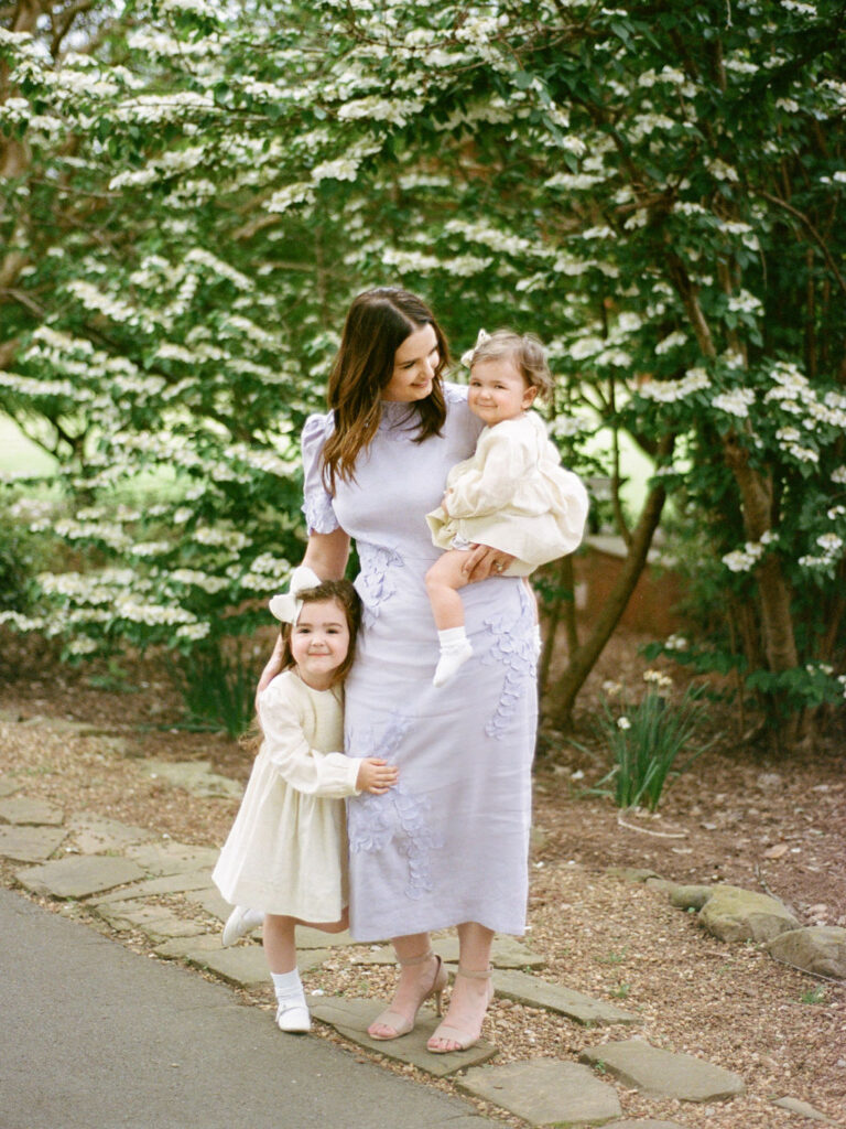 A mother poses, at a Huntsville Botanical Garden family photo session, with her two young daughters, one held on her hip and the other standing next to them