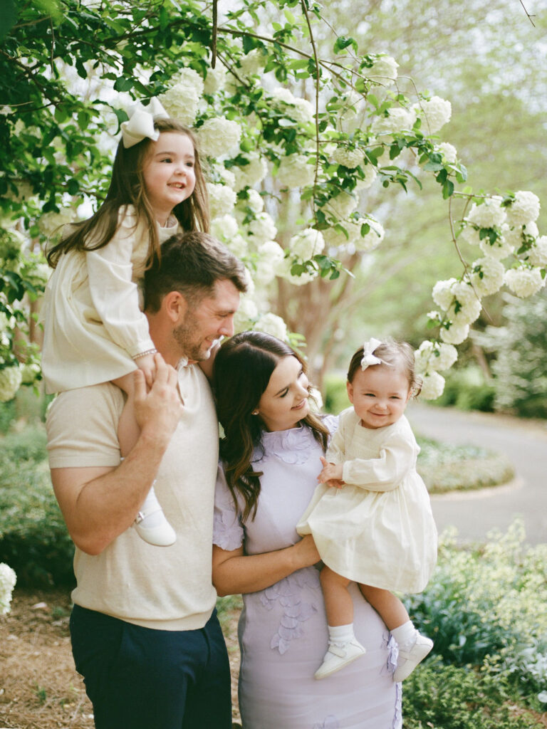 A family of four posing in the Huntsville Botanical Garden, with one toddler on dad's shoulders and the other in mom's arms, while they all laugh. 