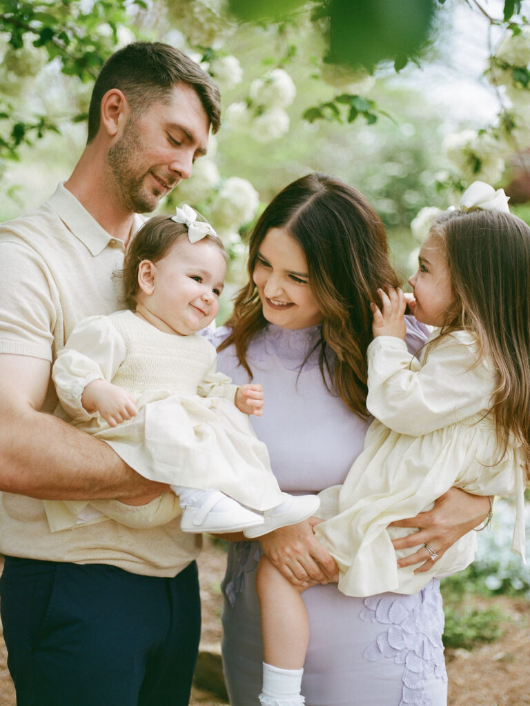 A candid moment where a family of four laughs together in the Huntsville Botanical Garden, with one toddler held by each parent.