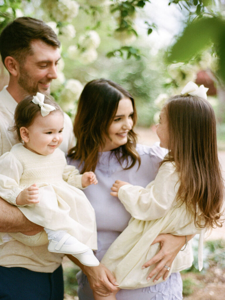 A sweet moment where a family of four smiles at each other in the Huntsville Botanical Garden, with one toddler held by each parent.