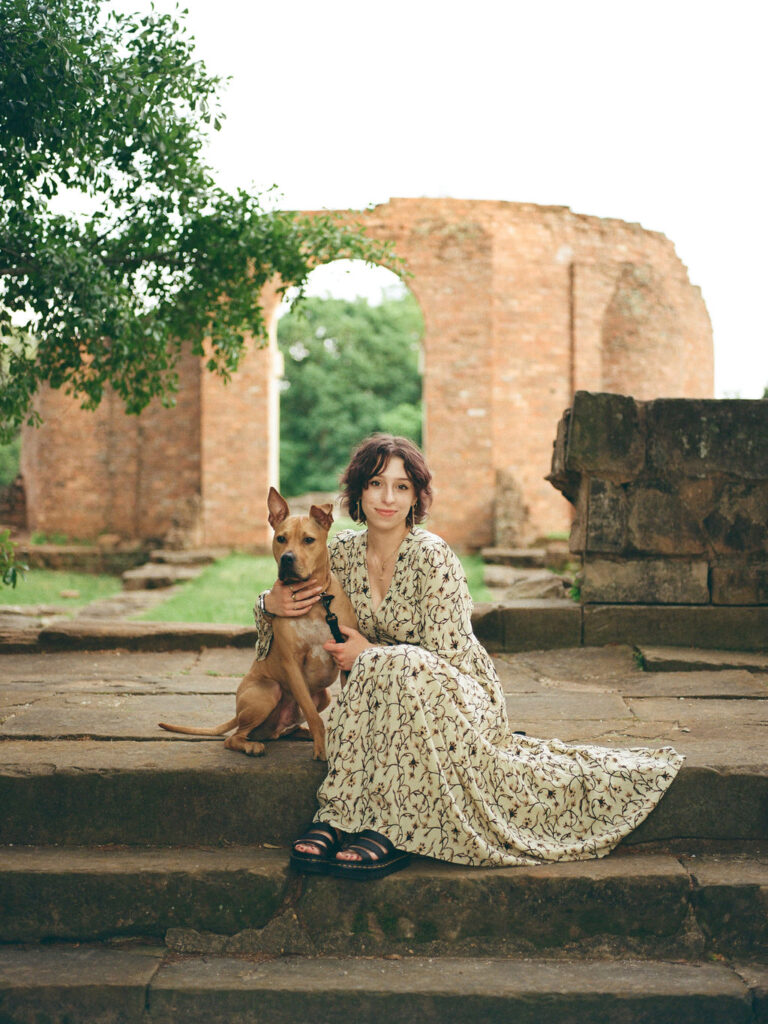 A woman in a floral Doen dress sits on stone steps, looking at the camera, with her medium-sized, brown dog,