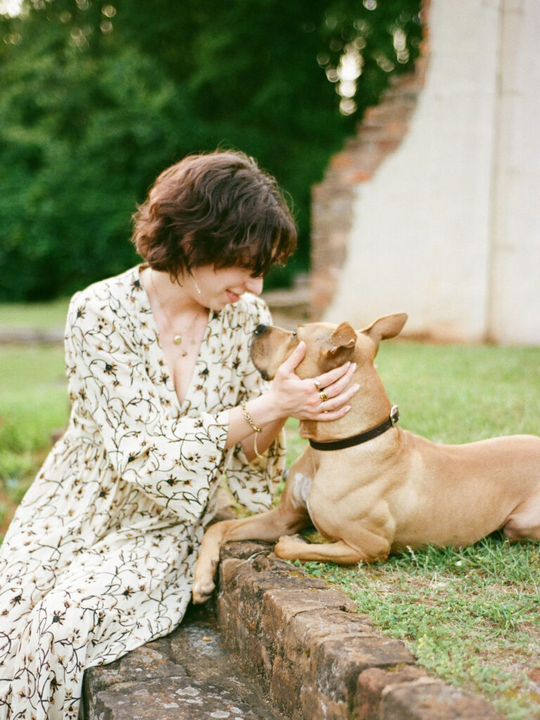 A woman in a floral Doen dress sits  with her brown pitbull/catahoula mix during their pet and owner photoshoot with a Huntsville pet photographer.