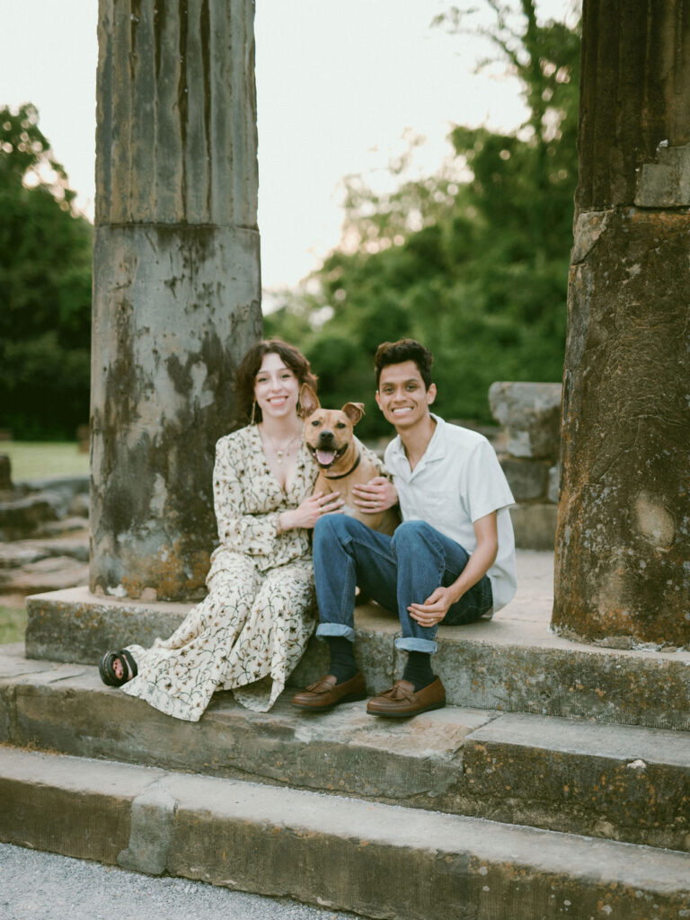 A couple and their dog, sitting on stone steps, all smile at the camera during their dog and owner photoshoot.