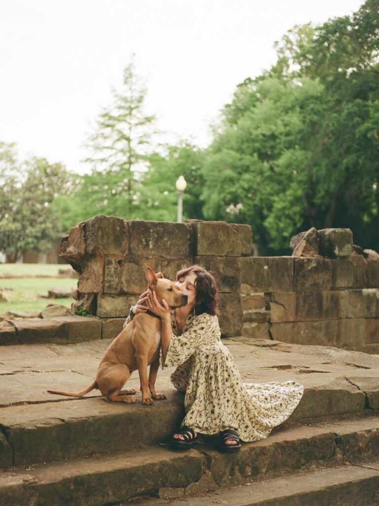 A woman in a floral Doen dress sits on stone steps with her medium-sized, brown dog and kisses his cheek.