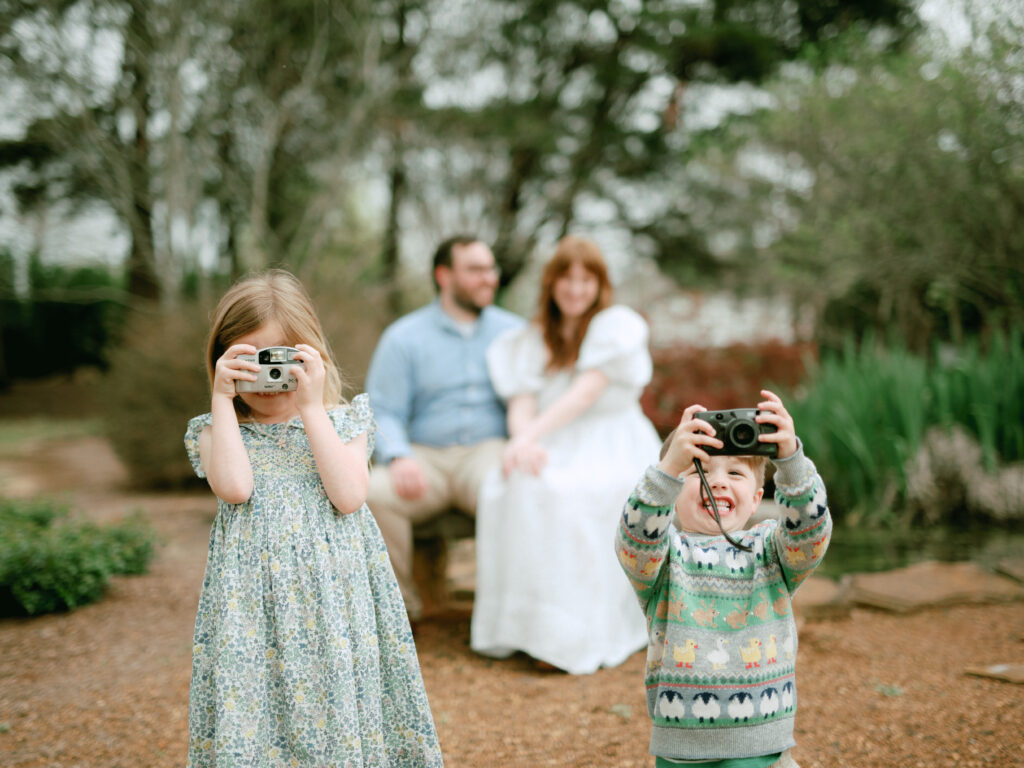 Two small children face the camera holding point-and-shoot cameras, in the foreground, while their parents sit in the background