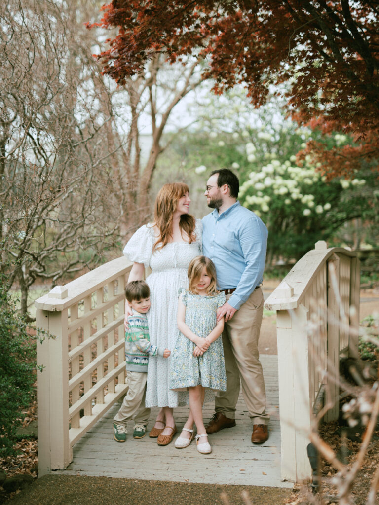 A family, two adults and two young children, pose together on a small wooden bridge at the best photoshoot location in Huntsville, the Huntsville Botanical Garden.
