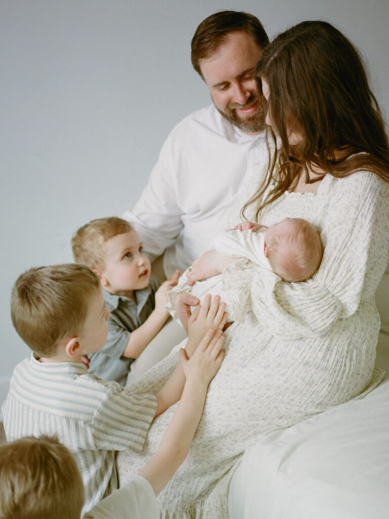 Parents sit on a bed, the mom holds their newborn baby son, while their three older children sit on the floor looking up at their baby brother