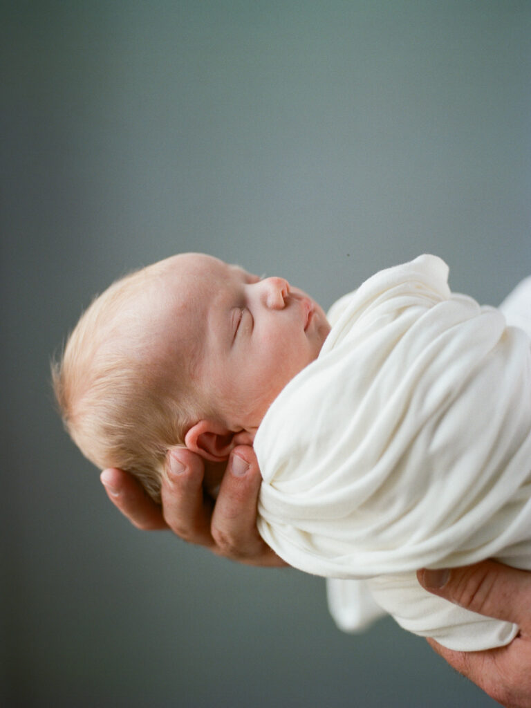 Closeup of a newborn baby in a white swaddle held by his father during their session with Huntsville maternity and newborn photographer Fauna & Film