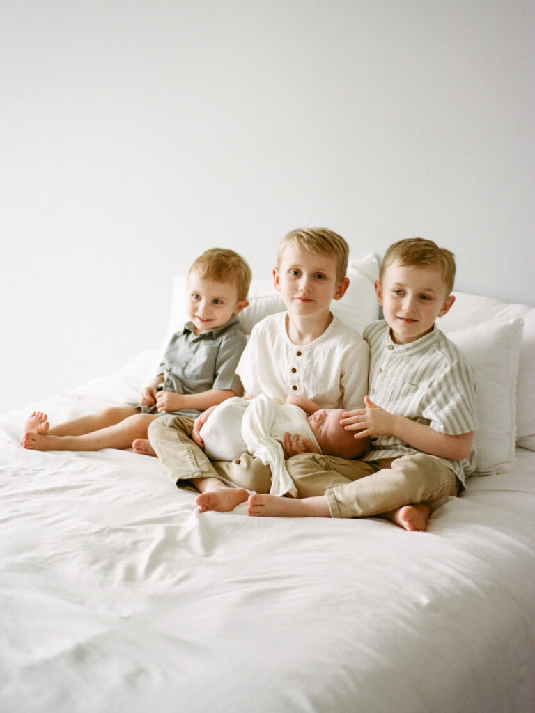 A newborn baby in a white swaddle held by his brother while their other two brothers sit next to them
