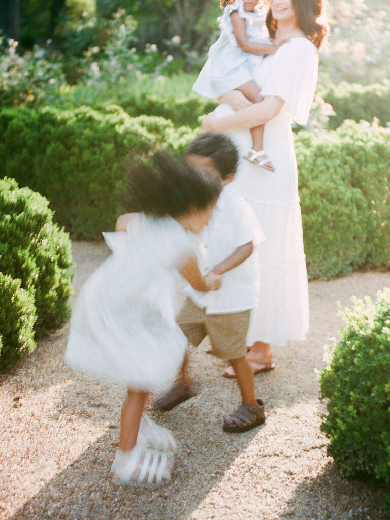 Twin four-year-olds play ring around the rosie while their mom, holding her youngest child, watches and smiles