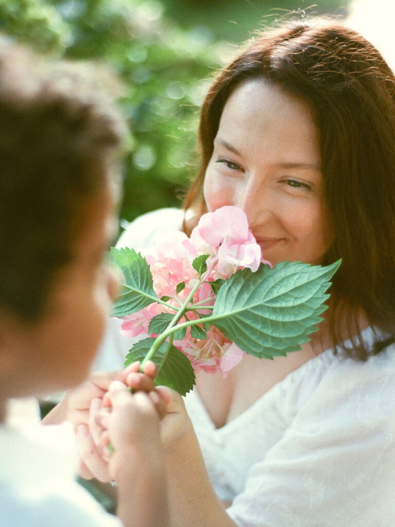 A mom smiles as she smells the pink hydrangea offered to her by her son