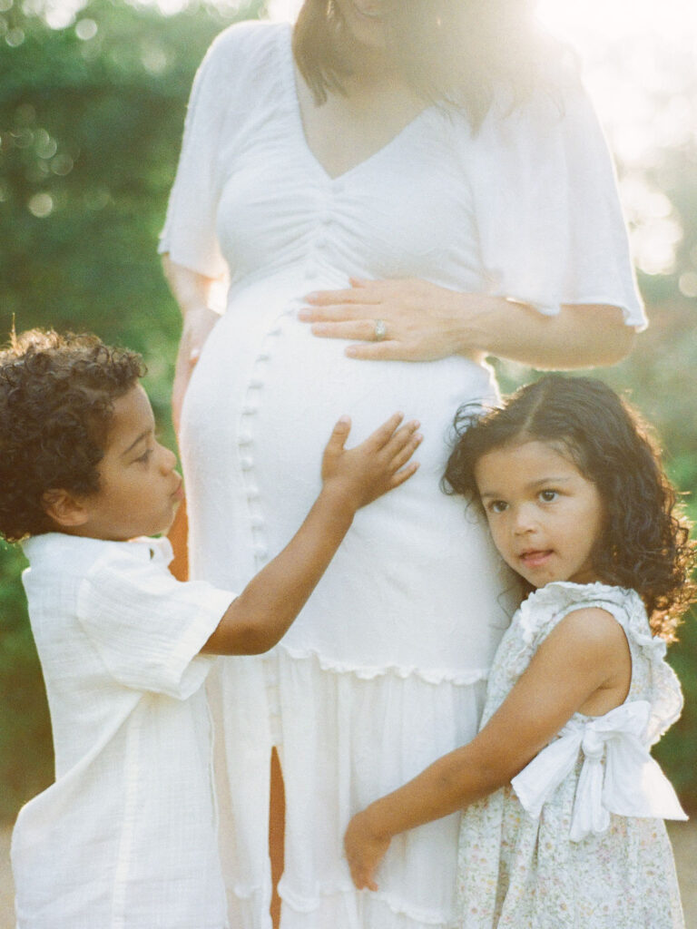 Twin four-year-olds pose with their heavily pregnant mom. One child hugs her mom's leg while the other child kisses his mom's baby bump.