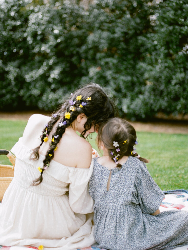 A woman and her young daughter sit on a picnic blanket, facing away from the camera, showing their hair braided with flowers