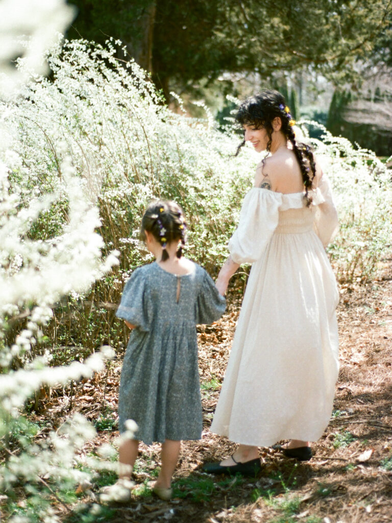 A mother and her young daughter, both wearing flowy dresses with flowers in their hair, hold hands as they admire a garden of flowers.