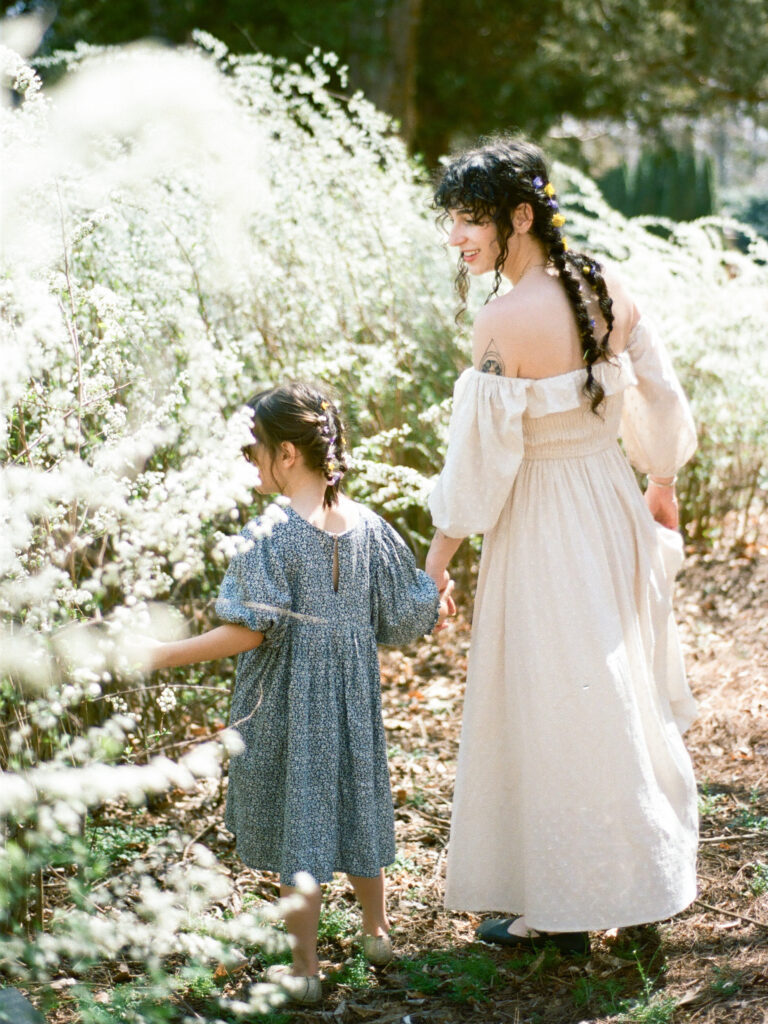 A mother and her young daughter, both wearing flowy dresses with flowers in their hair, hold hands as they admire a garden of flowers.
