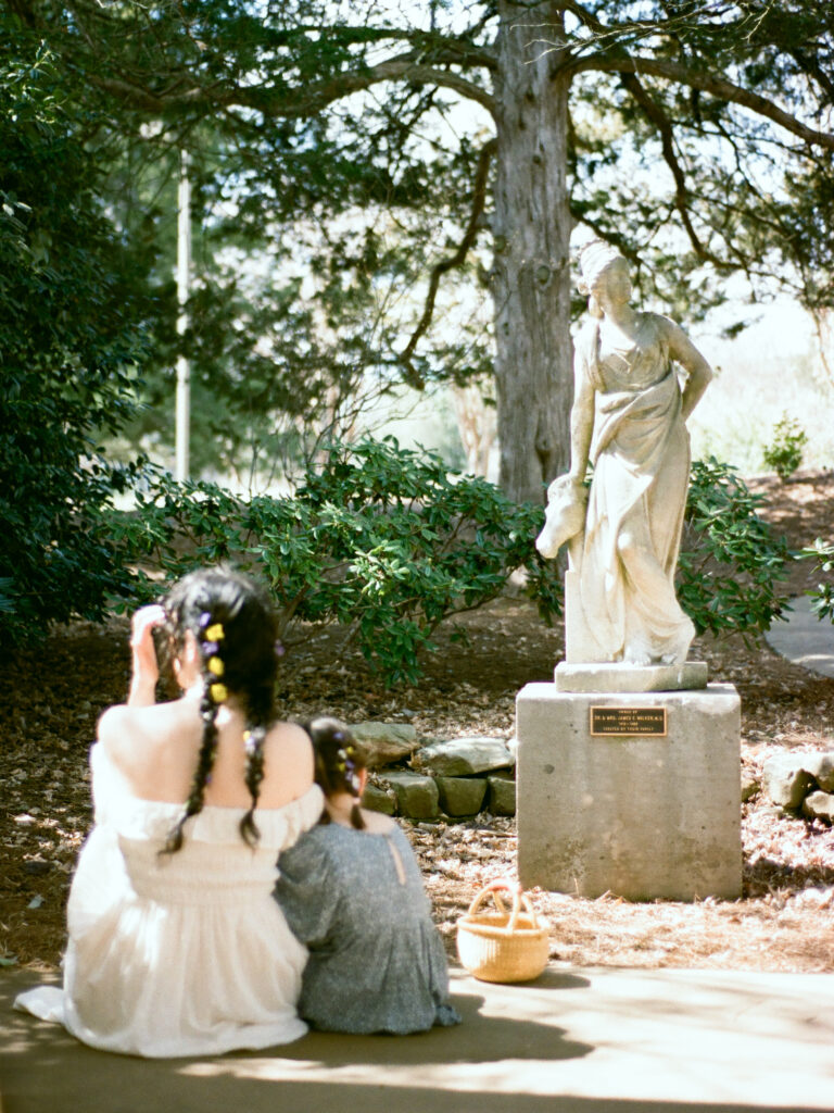 A mother and her young daughter sit on the ground and look at a statue of a woman