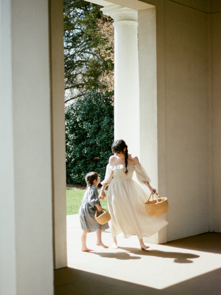 A woman in a white dress and her young daughter in a blue dress, both holding baskets, hold hands and look at each other, at their Huntsville motherhood photography session