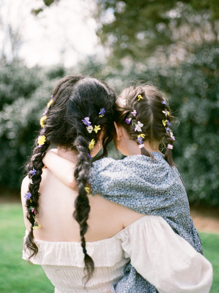 A mother stands holding her daughter, facing away from the camera. Both of them have flowers in their braided hair.