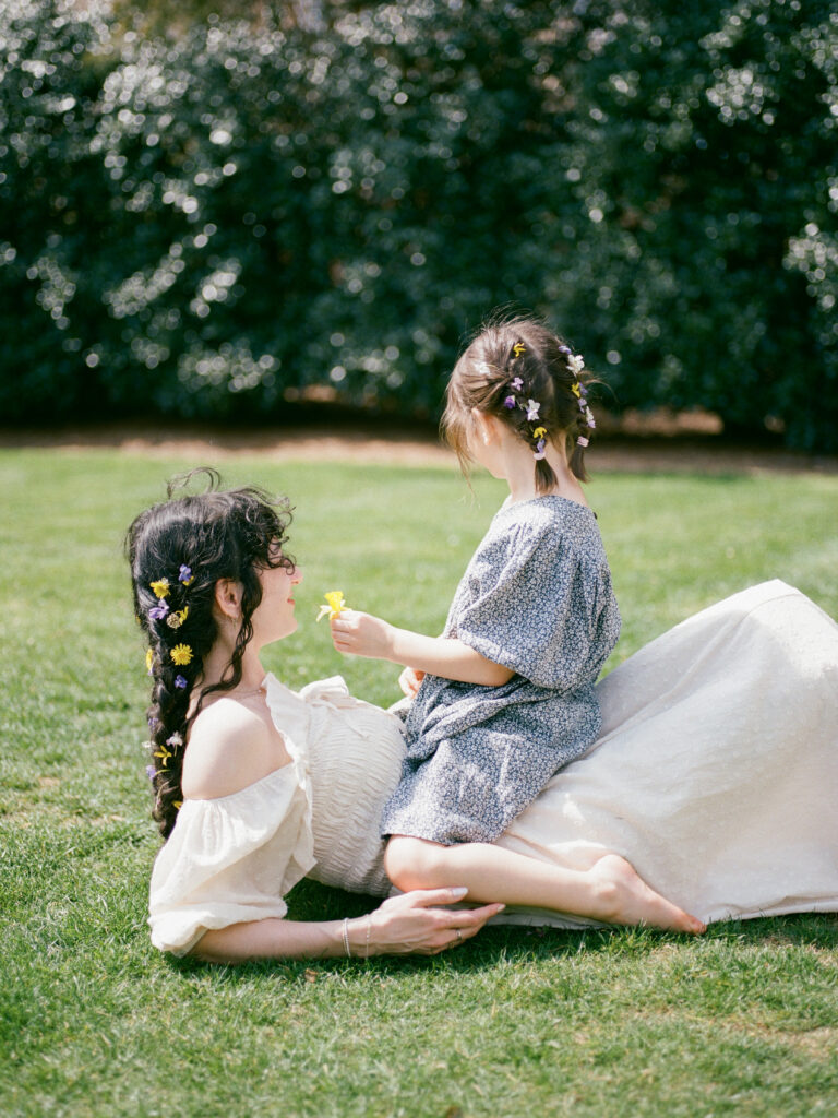 A woman lies in a grassy field with her small daughter sitting on her lap