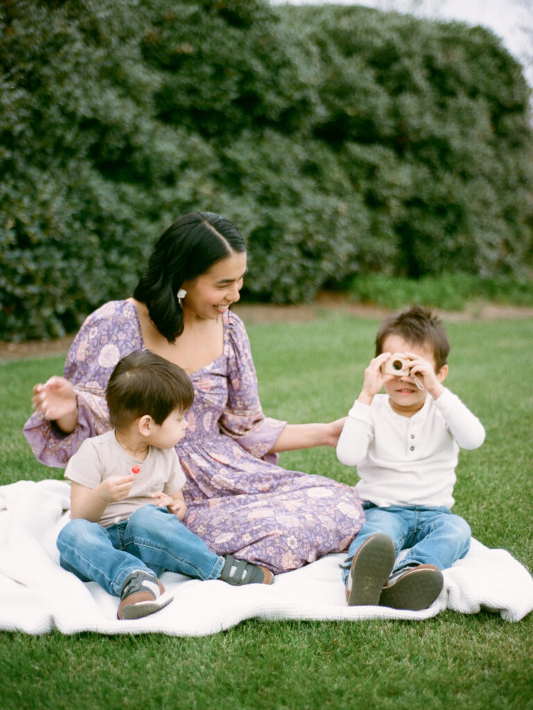 A woman in a purple dress sits on a picnic blanket with her two small children, one is holding a toy camera and the other is holding a red lollipop.