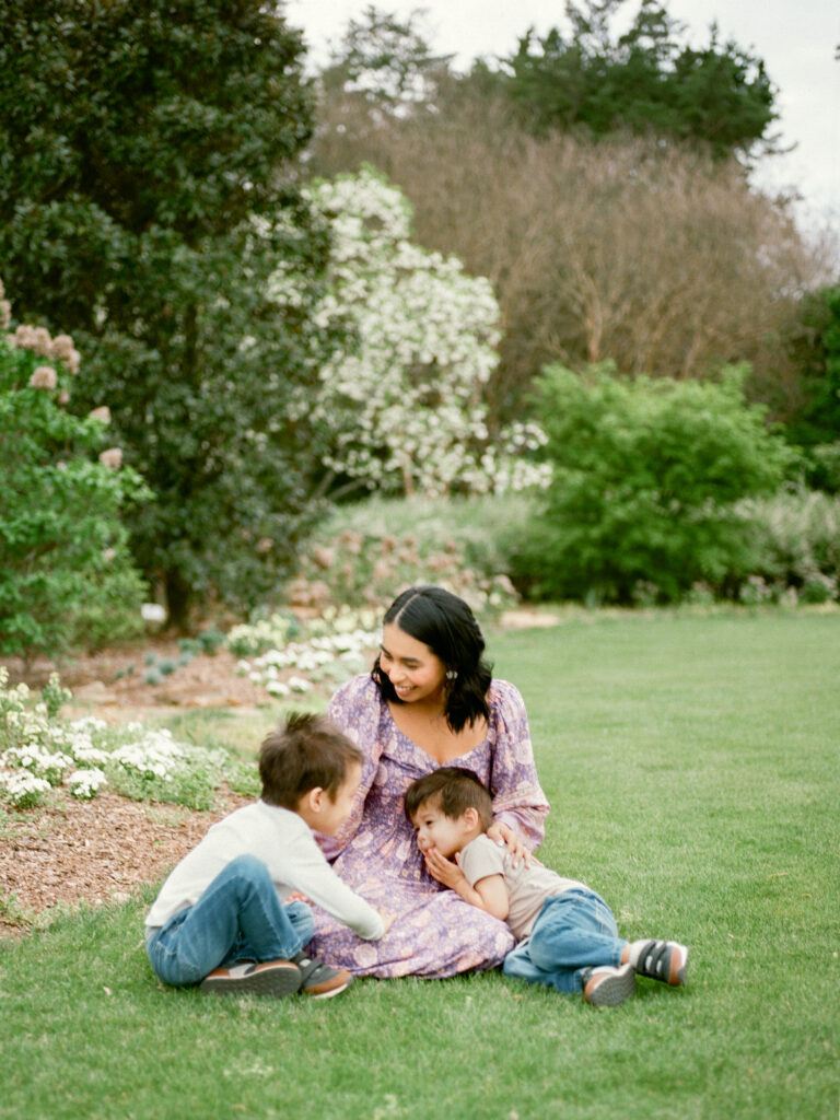 A woman in a purple dress sits in a lush green field with her two young children.
