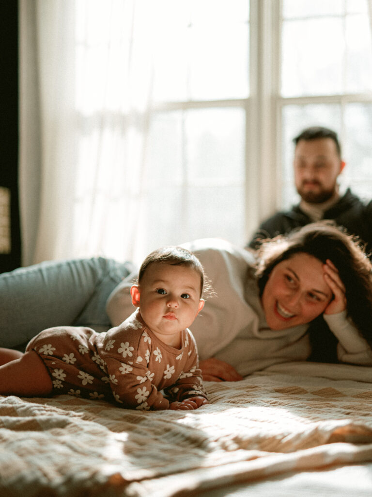 a baby lies down on a bed as her parents watch during their family photos at home