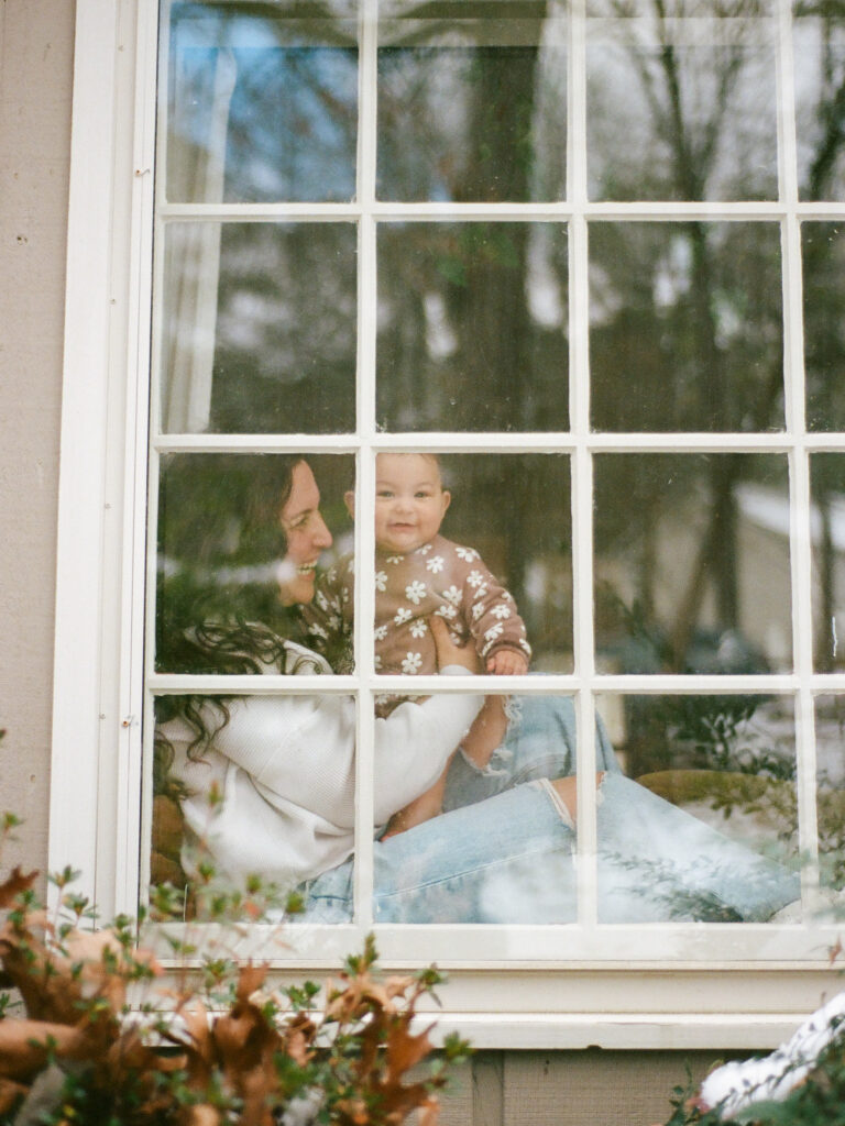 A mother and her baby seen through a window, smiling and laughing as they take family photos at home