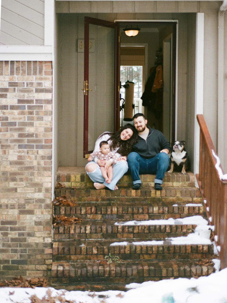 A man, a woman, a baby, and a dog sit together and smile on the front porch steps of their house during their family photos at home
